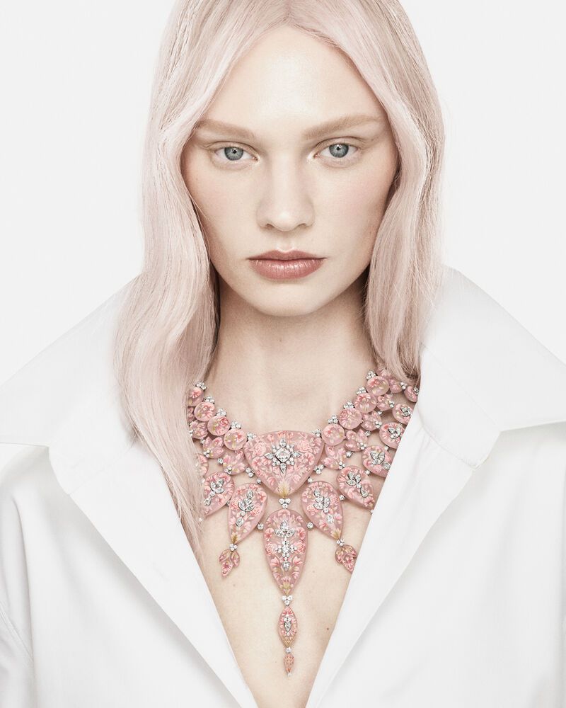 Model with platinum blonde hair wearing an ornate pink and diamond statement necklace and a crisp white collared shirt.