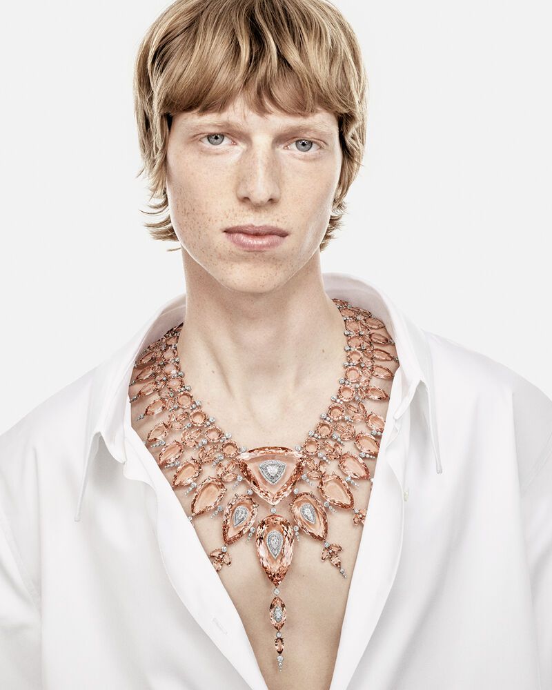 Close-up portrait of a male model wearing an elaborate gemstone necklace and unbuttoned white shirt against a white background