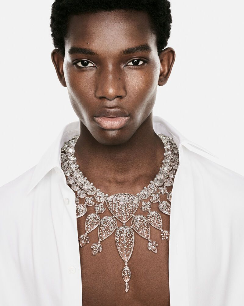 Portrait of a Black man wearing an elaborate diamond high jewelry necklace and an open white collared shirt against a white backdrop.