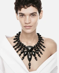 Model with short dark hair wearing an elaborate black statement necklace with diamond accents against a white studio background.