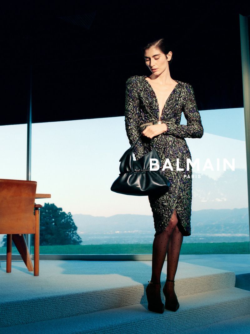 Fashion model in a textured long-sleeved midi dress holding a black leather handbag in front of panoramic glass windows.