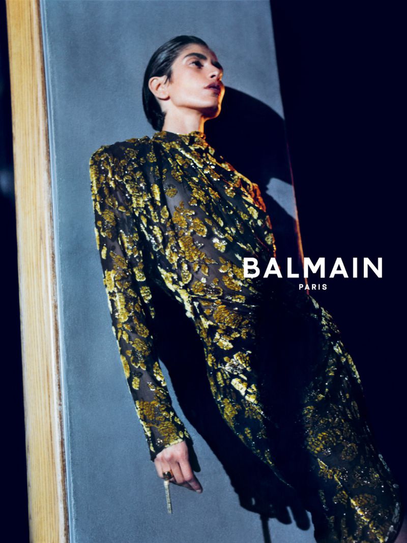 A model in a metallic gold and black long-sleeve dress poses against a grey panel with a bold shadow for Balmain.