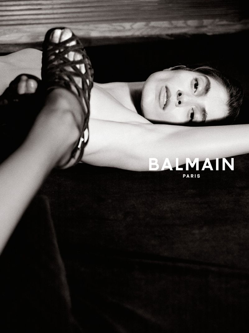 Black and white fashion campaign photograph of a woman reclining with a black strappy heeled shoe in the foreground and Balmain Paris logo.