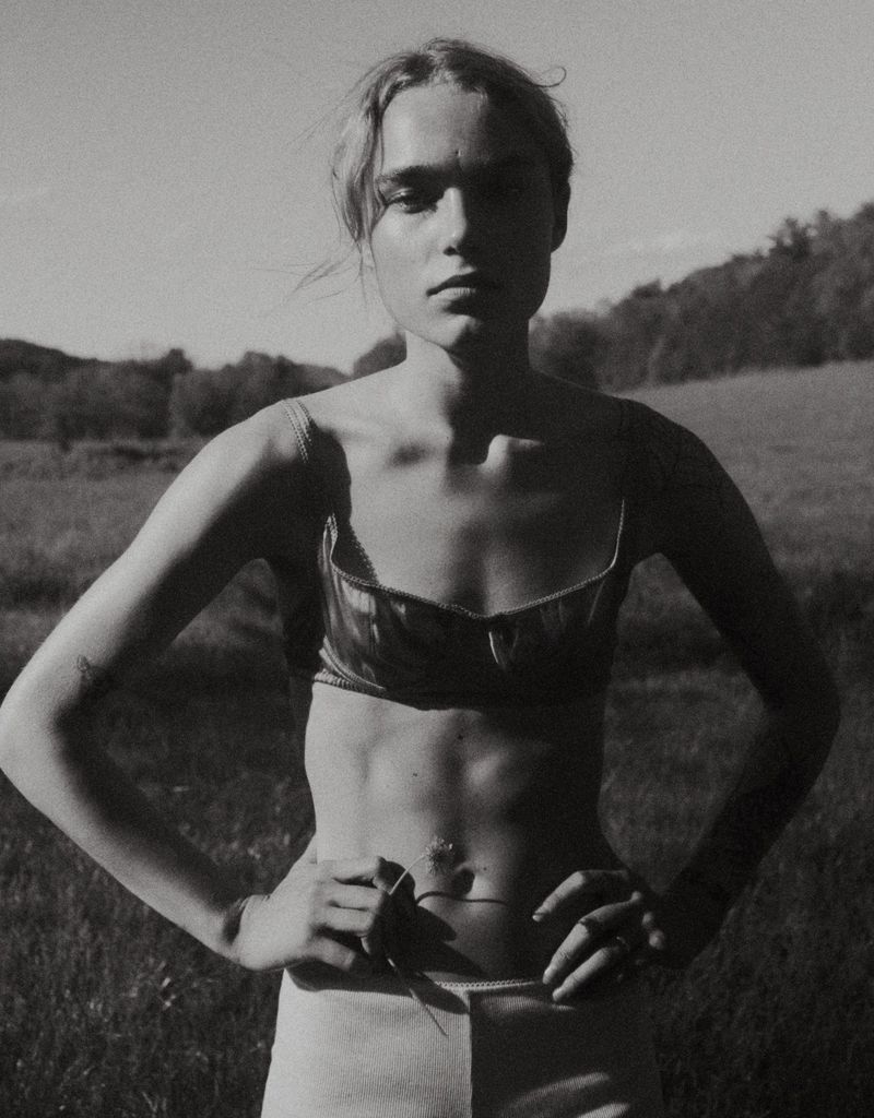 Monochrome portrait of a model in a satin bra top and light shorts standing with hands on hips in a grassy field.