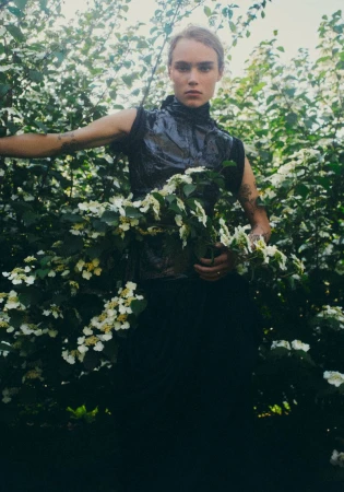 Model wearing a textured high-neck sleeveless black top posing amidst blooming white flowering bushes outdoors.
