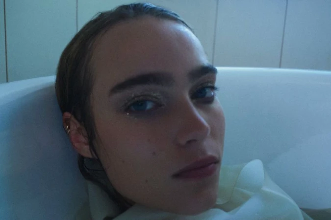 Close-up portrait of a woman with dark hair and glitter eye makeup resting her head back in a bathtub.