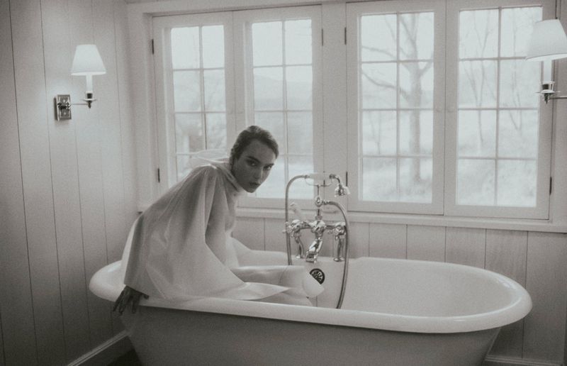 Woman in a sheer white draped garment seated inside a vintage freestanding bathtub in front of a window