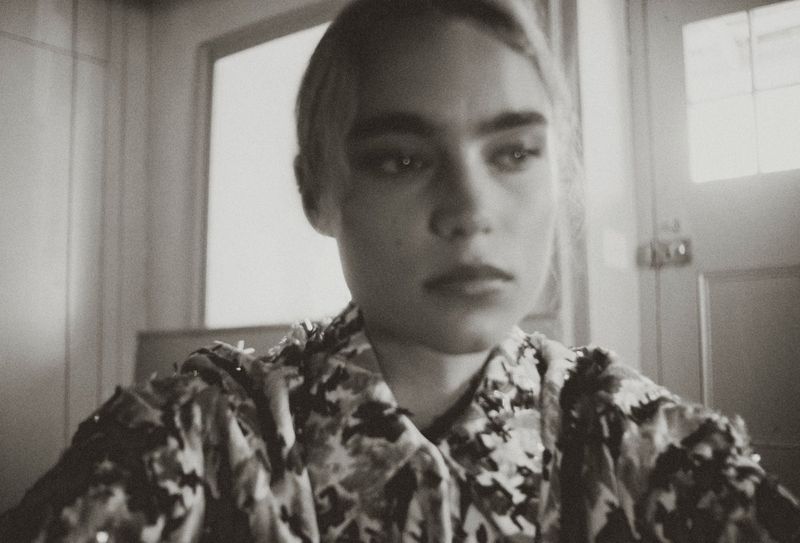 Monochrome portrait of a woman with dark eyebrows in a patterned jacket looking off-camera indoors near a windowed door.