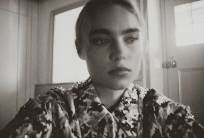 Monochrome portrait of a woman with dark eyebrows in a patterned jacket looking off-camera indoors near a windowed door.