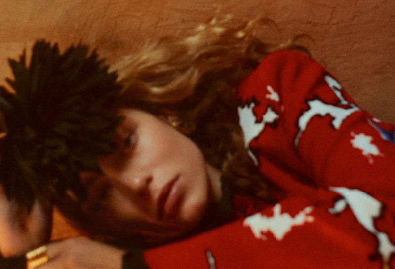 Model reclining in a red patterned knit sweater and spiky dark headpiece under warm atmospheric lighting.