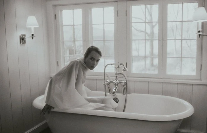 Woman in a sheer white draped garment seated inside a vintage freestanding bathtub in front of a window