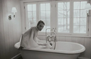 Woman in a sheer white draped garment seated inside a vintage freestanding bathtub in front of a window