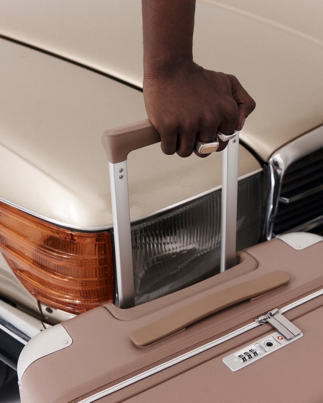 Close-up of a black man's hand holding a tan suitcase handle in front of an antique car with an amber headlight.
