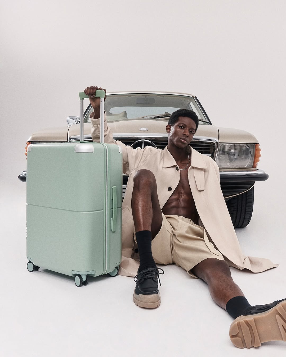 A black male model in a trench coat poses near a light-green rolling suitcase and classic car in a brightly lit studio setting.