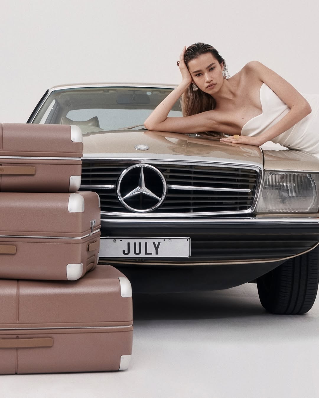 Model in white dress leaning on the hood of a vintage gold Mercedes next to stacked brown suitcases