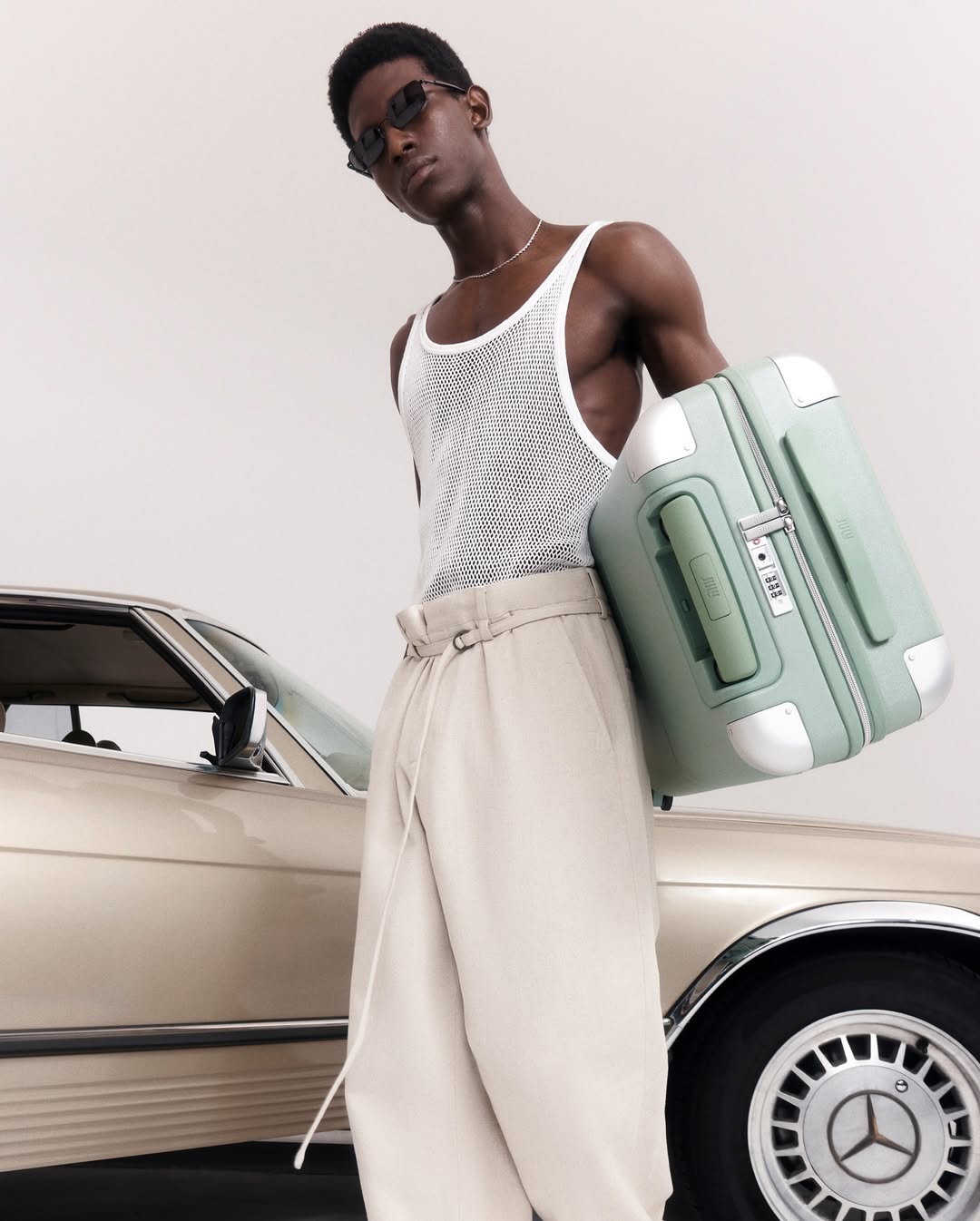 A fashionable man wearing sunglasses, a tank top, and pants poses with a vintage car and mint green suitcase in a studio shot.