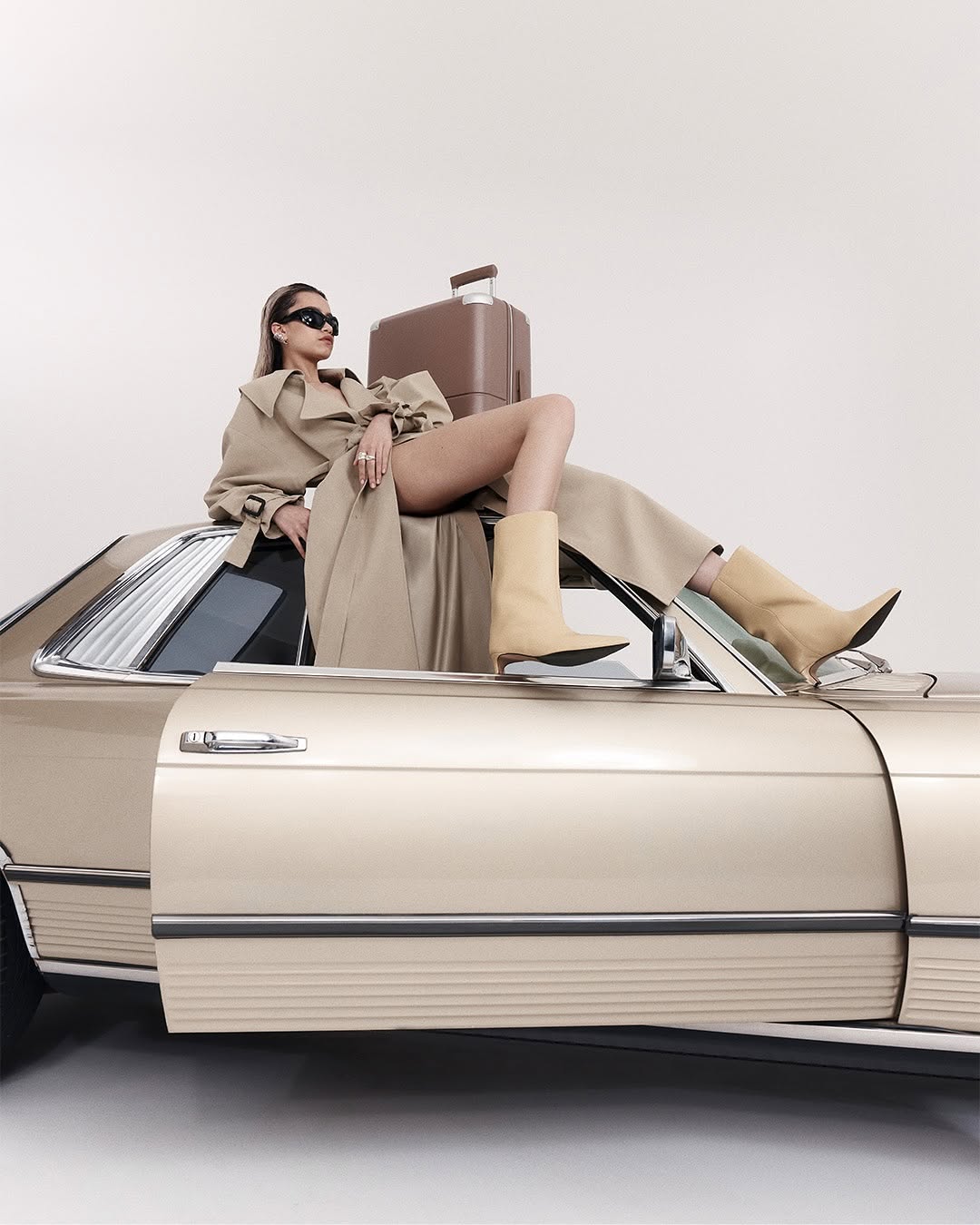 Model in beige trench coat and light boots reclining on a classic car roof with a brown suitcase against a white background