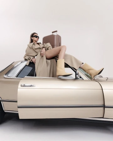 Model in beige trench coat and light boots reclining on a classic car roof with a brown suitcase against a white background