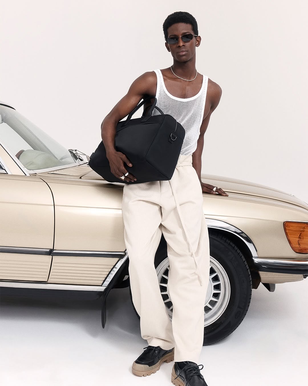 Man in tank top leans on classic car with black duffel bag. He wears sunglasses and beige pants against a white backdrop.