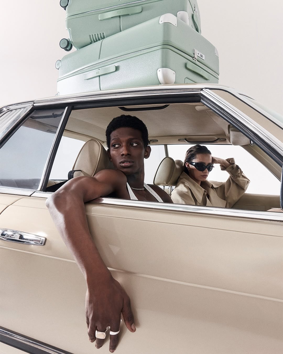 Two people in a vintage car with stacked luggage on top. The driver is a muscular black man, with a stylish woman in the passenger seat.