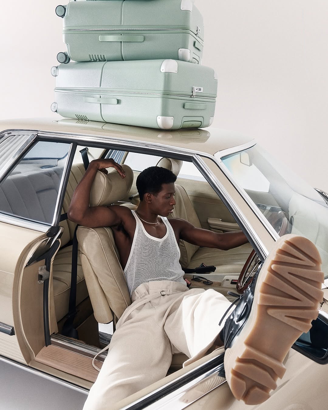 Stylish man poses in a vintage car with stacked luggage, showcasing fashion and travel aesthetics in a studio environment.