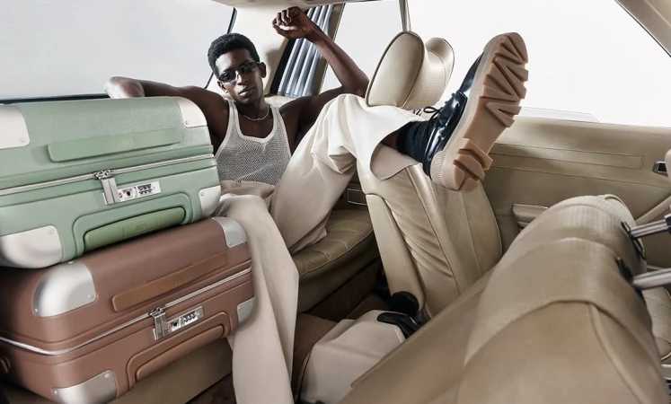 Stylish black man wearing sunglasses poses inside car with stacked suitcases in an indoor setting, capturing a fashion and travel aesthetic.