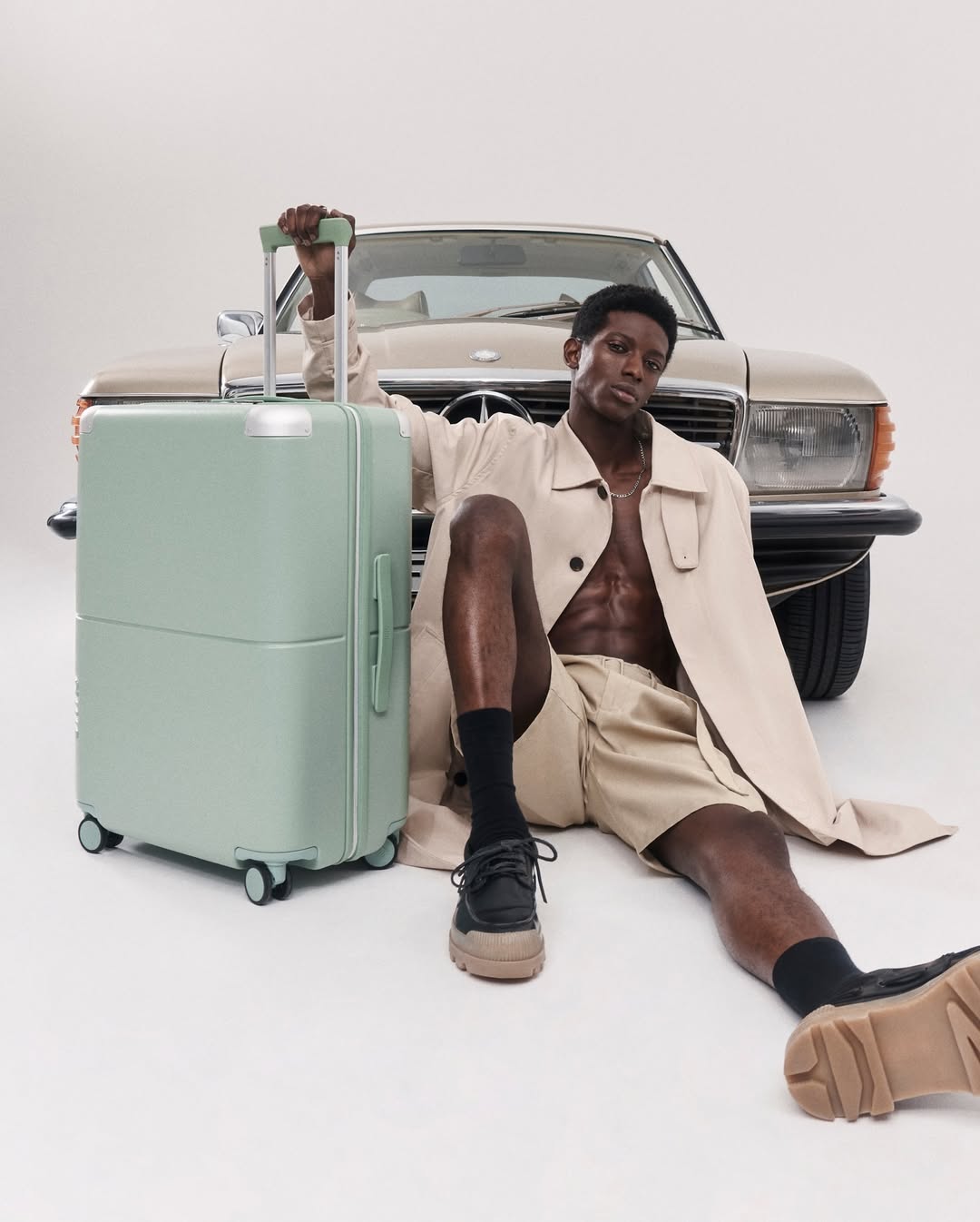A man poses with a mint-green suitcase and a beige vintage car in a studio. He is wearing beige shorts and a trench coat.