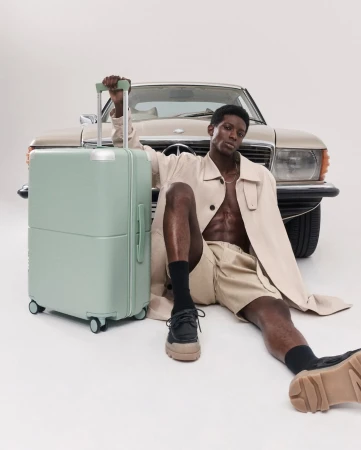 A man poses with a mint-green suitcase and a beige vintage car in a studio. He is wearing beige shorts and a trench coat.