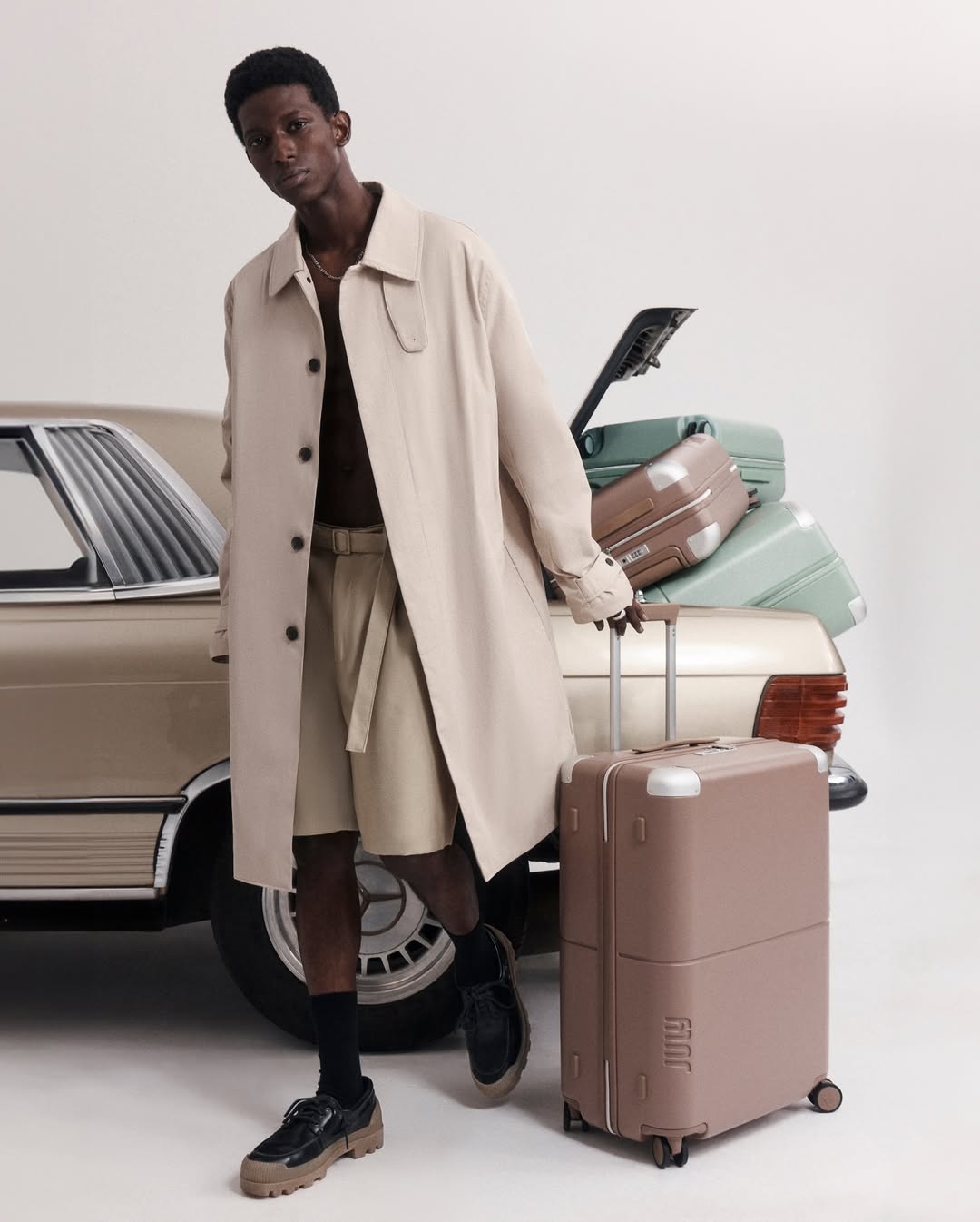 Dark-skinned adult male poses next to vintage car and luggage set in neutral tones, wearing belted trench coat and shorts in a studio shot.