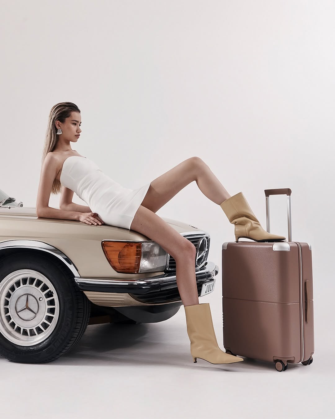 A woman in a white dress and beige boots poses on a beige vintage car next to a brown suitcase in a clean, well-lit photo studio.