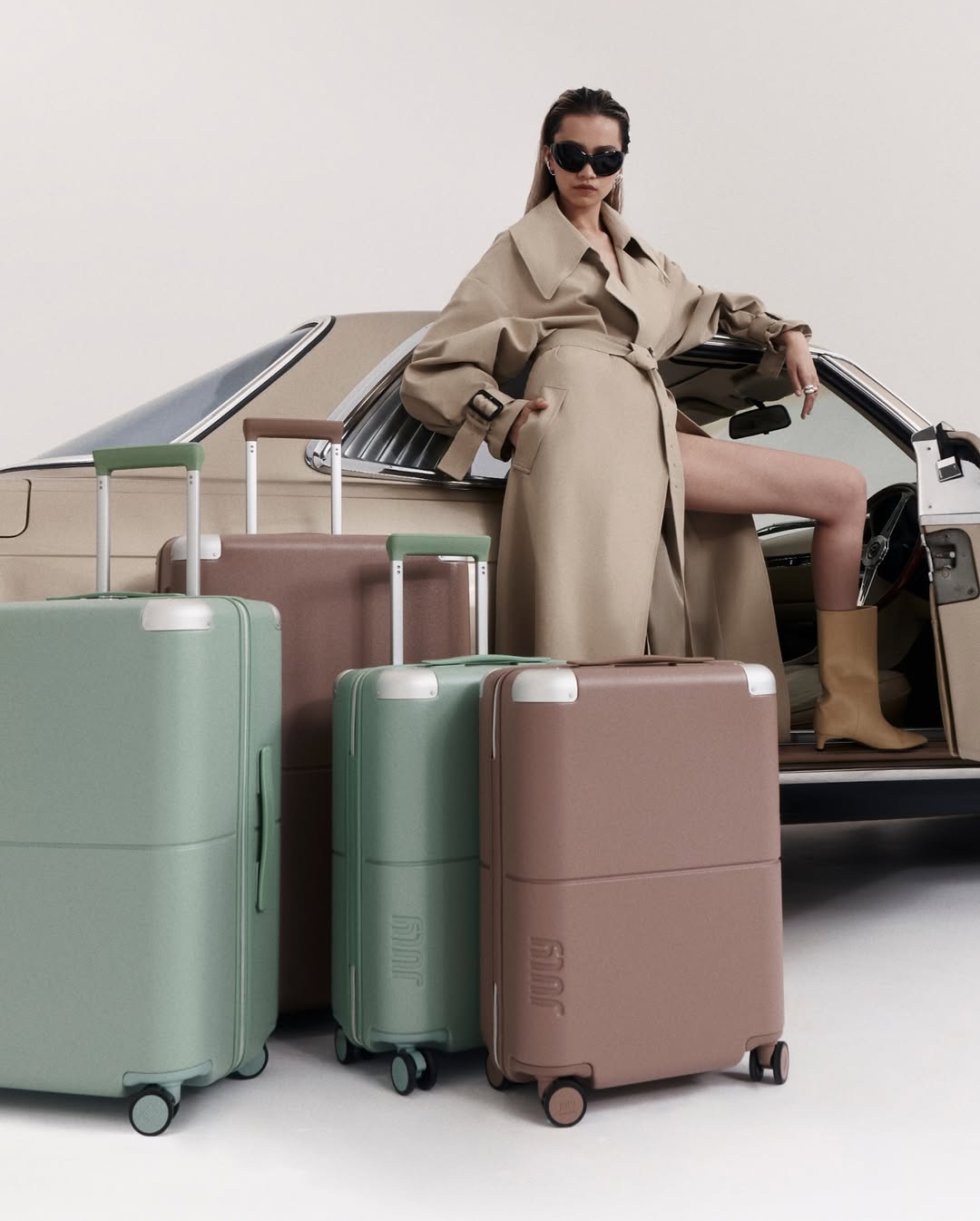 Fashionable woman in trench coat poses by vintage car with collection of pastel rolling luggage in studio setting. 