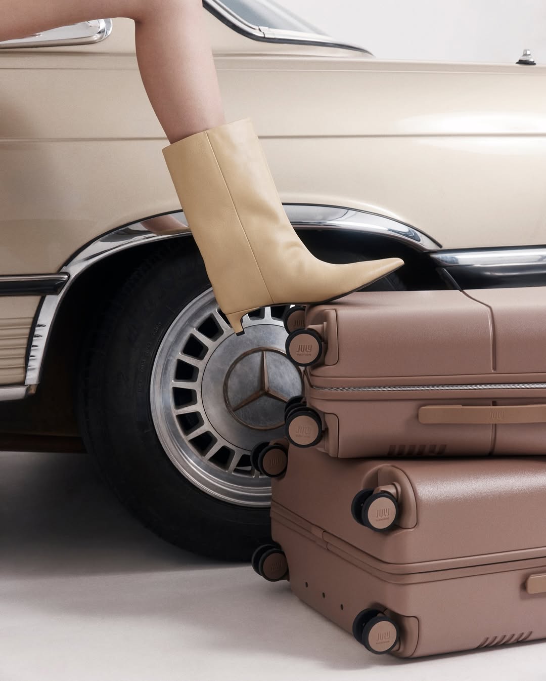 Woman's leg with tan pointed boot resting on a stack of suitcases, with a portion of vintage car in background.