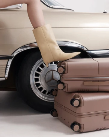 Woman's leg with tan pointed boot resting on a stack of suitcases, with a portion of vintage car in background.