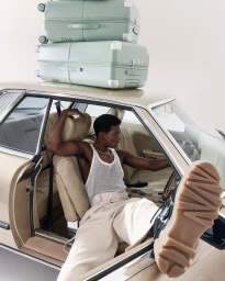 A Black man wearing a white mesh tank top seated inside a classic car with green suitcases stacked on top.