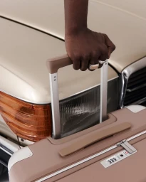 Close-up of a person holding the handle of a taupe suitcase with a vintage car and another suitcase in the background.