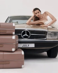 Model in white dress leaning on the hood of a vintage gold Mercedes next to stacked brown suitcases