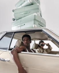 A man and woman sit inside a beige car with a tall stack of sage green suitcases loaded on the roof.