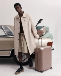 Black man standing beside a vintage car wearing a beige coat and holding a pink rolling suitcase