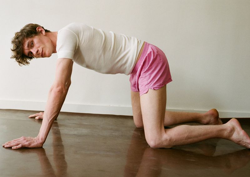 Adult male in a bent-over yoga position in a light indoor studio. He wears a white t-shirt and pink shorts on a shiny brown floor.
