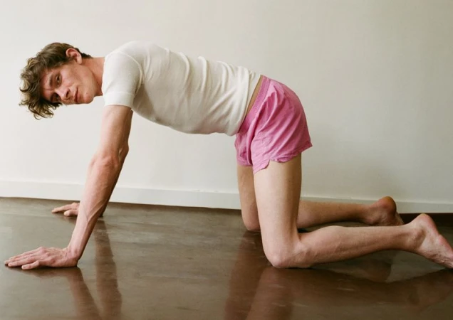 Adult male in a bent-over yoga position in a light indoor studio. He wears a white t-shirt and pink shorts on a shiny brown floor.