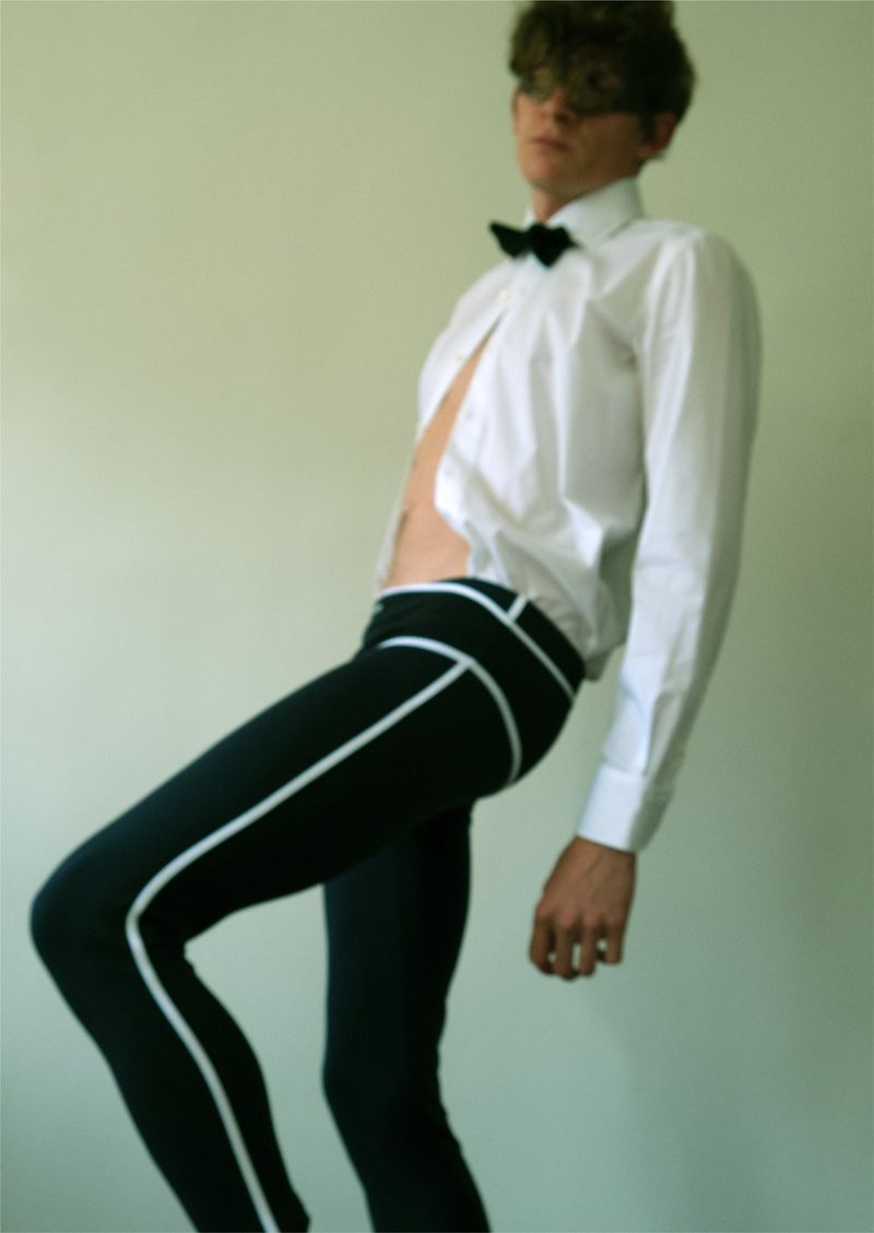 Stylish young adult male poses in a white shirt and black pants with white stripes, wearing a black bow tie against a plain background.