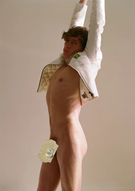Male model poses nude with a white jacket, shot in a photo studio with neutral background. The image has studio lighting.