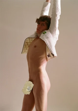 Male model poses nude with a white jacket, shot in a photo studio with neutral background. The image has studio lighting.