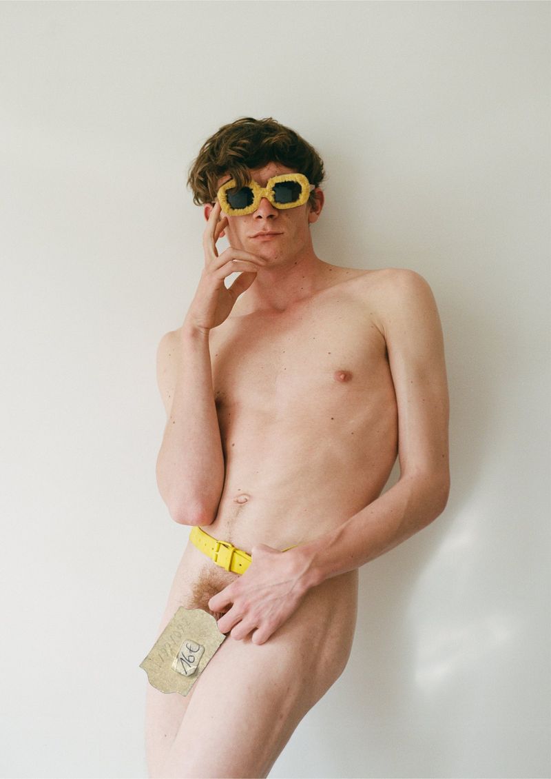 Nude adult male posing in front of white wall wearing yellow sunglasses and a belt with a piece of paper covering the groin.