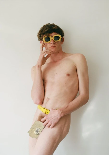 Nude adult male posing in front of white wall wearing yellow sunglasses and a belt with a piece of paper covering the groin.