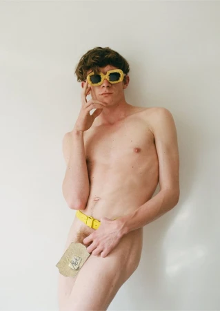 Nude adult male posing in front of white wall wearing yellow sunglasses and a belt with a piece of paper covering the groin.