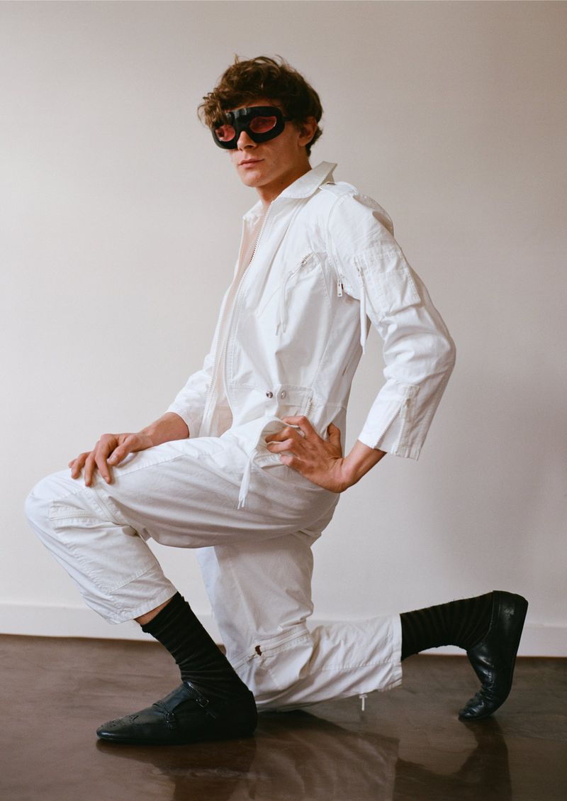 A person wearing a white jumpsuit, black socks, and black shoes kneels in front of a white wall indoors, highlighted by studio lighting.