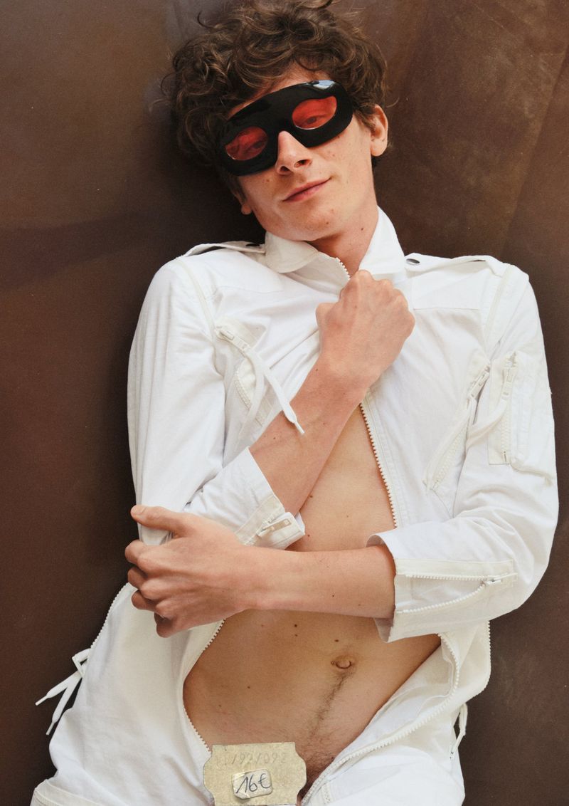 A light-skinned adult male with curly brown hair wearing a white one-piece and black eye mask, posing on a brown surface.