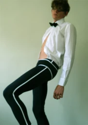 Young man in a white dress shirt with black bow tie and black trousers standing against a pale wall.