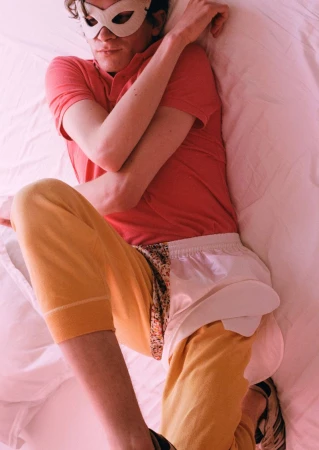 Person lying on white bed sheets wearing a white masquerade eye mask, coral polo shirt, and yellow leggings.
